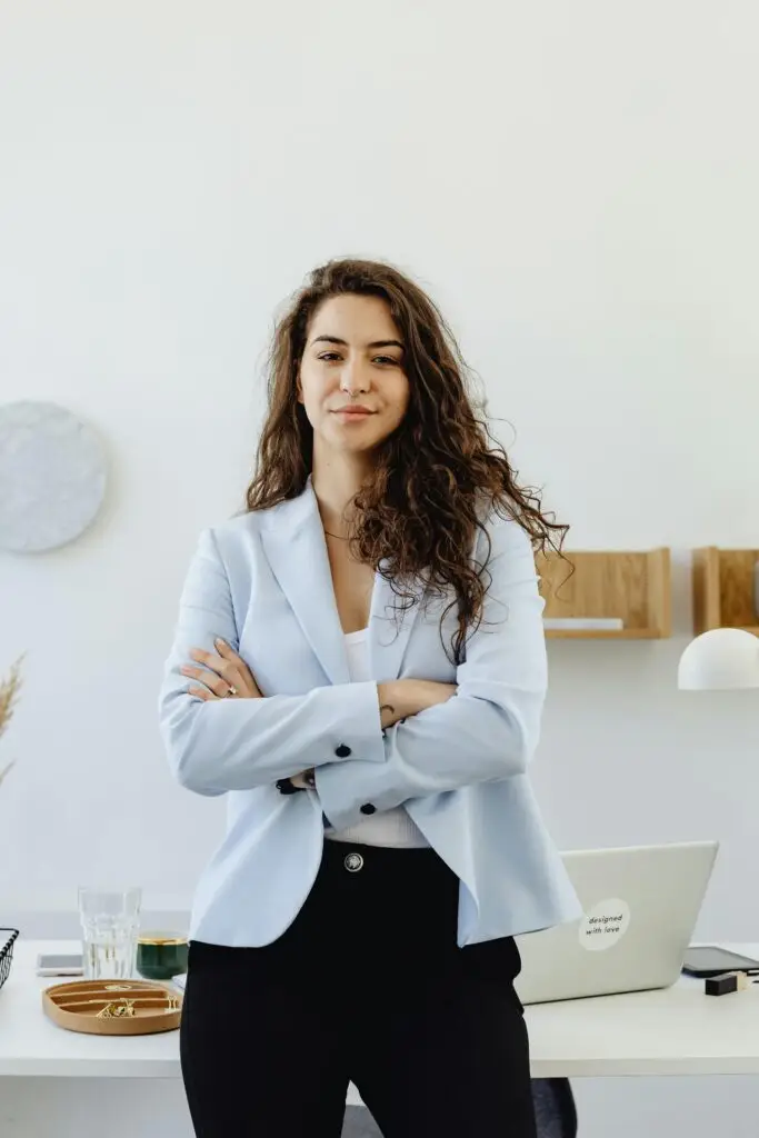 Professional woman in a white blazer standing confidently in an office with arms crossed.